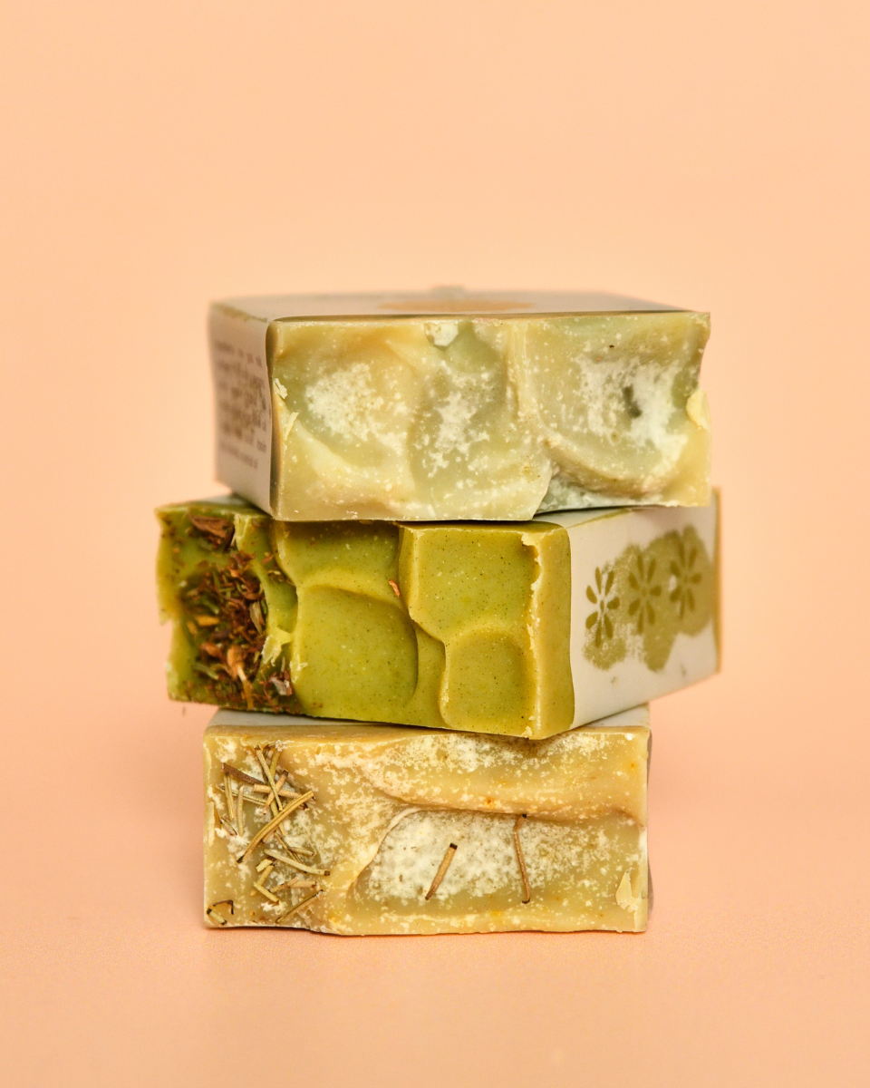 Three bars of goat milk soap with a golden hue and floral patterns on top, stacked on a pink background.